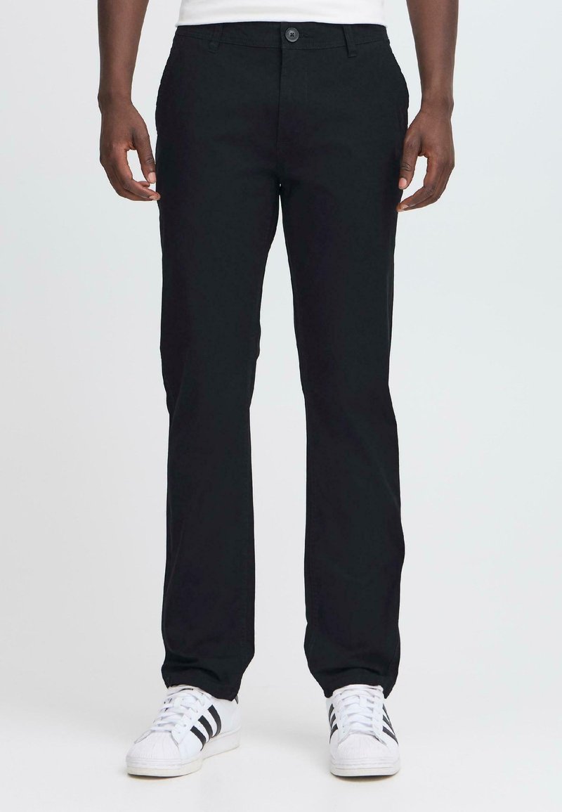 Homme portant un pantalon noir à jambes droites et des baskets blanches avec des rayures noires, se tenant devant un fond clair uni.