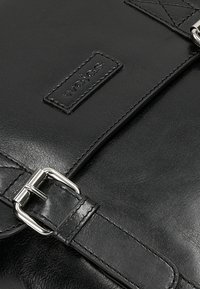 Borsa nera in pelle con superficie testurizzata, hardware argentato e patch logo rettangolare. Include tracolla regolabile e dettaglio della fibbia.
