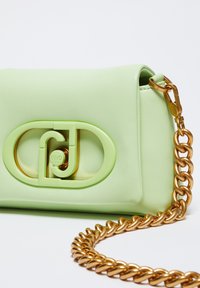 Borsa a tracolla in pelle verde chiaro con un logo circolare verde e oro ben visibile, dotata di una catena dorata spessa.
