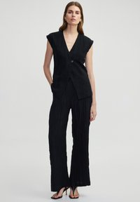Schwarzer ärmelloser Jumpsuit mit tiefem V-Ausschnitt, strukturierter Stoff, sichtbare Knopfdetails und weit geschnittene Hosenbeine, kombiniert mit offenen Zehenhighheels.