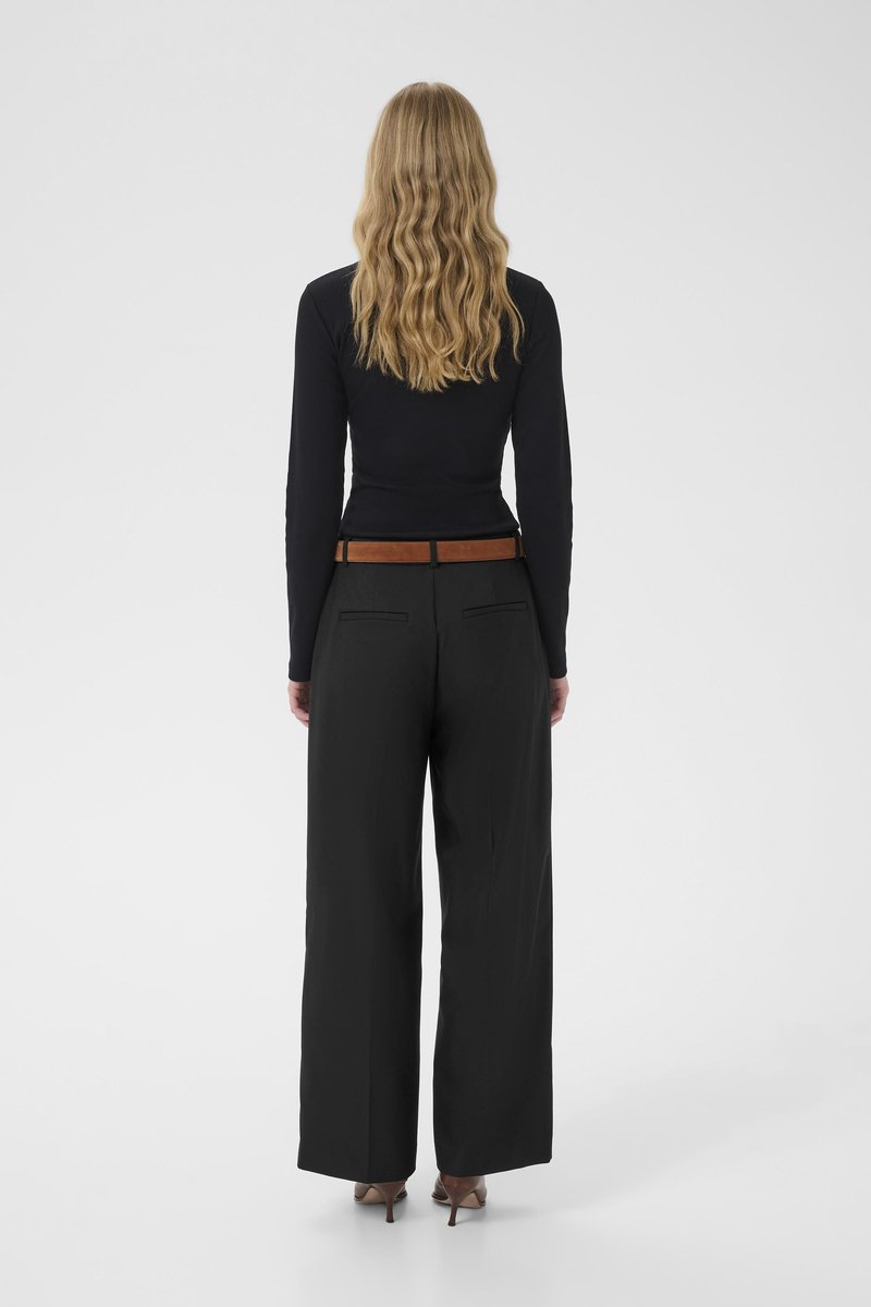 Haut noir à manches longues associé à un pantalon large noir taille haute. Ceinture en cuir marron ; chaussures à talons marron clair. Cheveux texturés tombant en cascade.