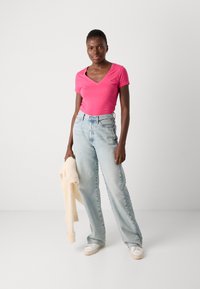 Roze v-hals t-shirt gecombineerd met lichtblauwe high-waisted jeans. De persoon houdt een crèmekleurige trui vast en draagt witte sneakers. Eenvoudig ontwerp, casual uitstraling.