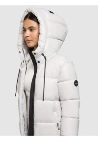 Bijela podstavljena jakna s visokim ovratnikom i velikim kapuljačom. Ima crne naglaske, zatvarač i vidljive šavove. Prikladna za hladno vrijeme.