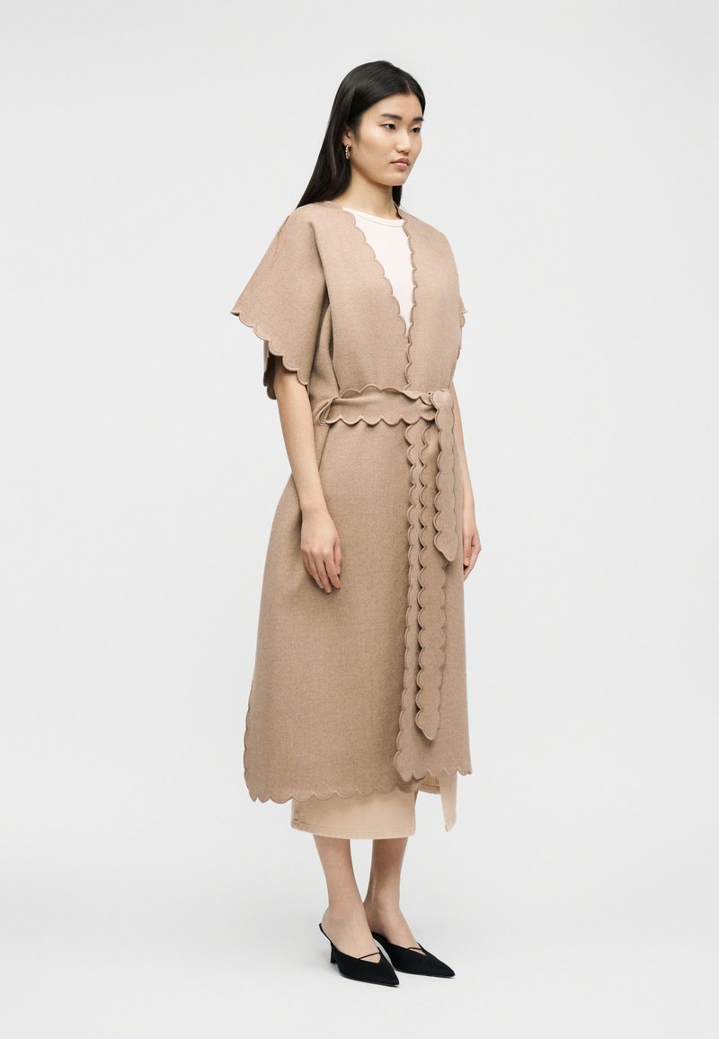 Frau trägt einen beigen, knieumspielenden Strickmantel mit gewellten Kanten, der in der Taille gebunden ist, kombiniert mit schwarzen spitzen Pumps.