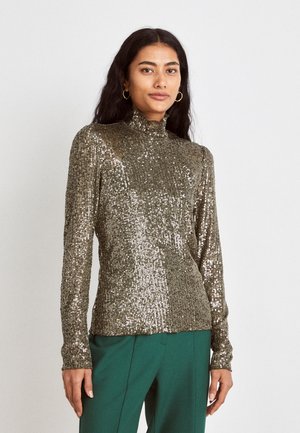 Frau mit langen dunklen Haaren, die einen goldenen Pailletten-Rollkragenpullover und grüne Hosen trägt, steht vor einem schlichten hellen Hintergrund.