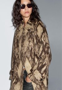 Brauner und beiger gemusterter Wollmantel mit Kragen, Knopfverschluss und umgeschlagenen Manschetten, kombiniert mit dunkler Sonnenbrille und einem schlichten Oberteil.