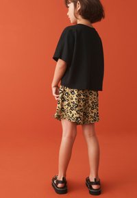 Sort kortærmet skjorte parret med en gul nederdel med sort leopardprint, afsluttet med en flæset kant, og sorte klumpede sandaler.