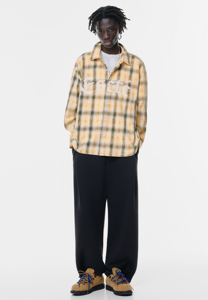 Camicia oversize a quadri giallo e verde con un logo bianco, pantaloni neri a gamba larga e scarpe beige con lacci blu e suole nere.