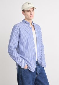 Blue and white striped button-up shirt with a classic collar, long sleeves, and a relaxed fit, paired with blue denim jeans and a beige cap.