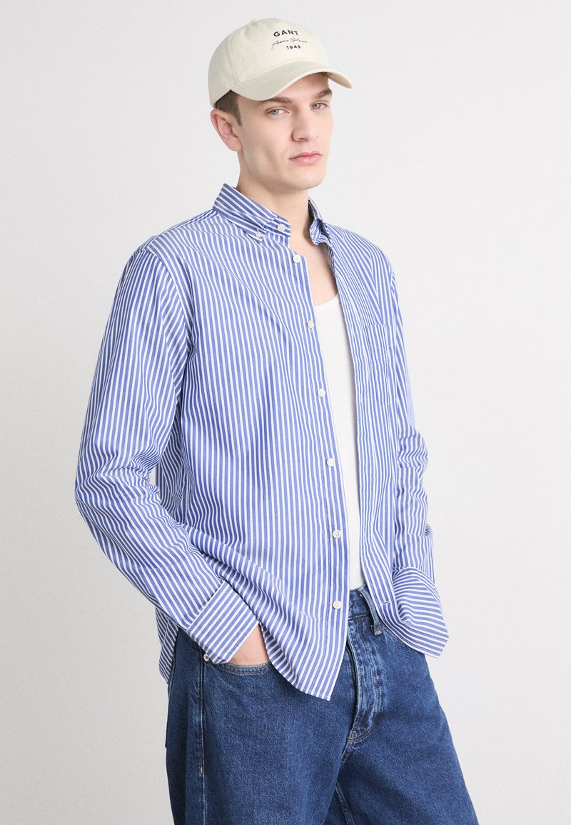 Blau-weiß gestreifte Knopf-bluse mit klassischem Kragen, langen Ärmeln und lockerer Passform, kombiniert mit blauen Jeans und einer beigen Kappe.