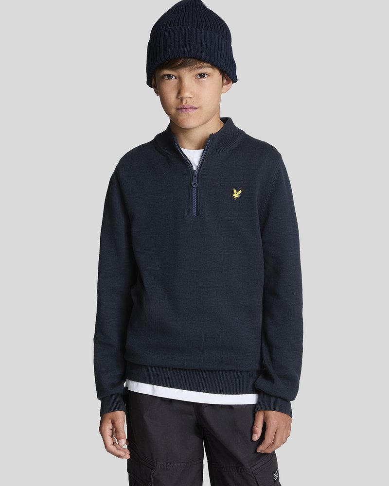 Cache-cœur bleu marine zippé avec des poignets et un ourlet côtelés, arborant un petit logo jaune. Associé à un bonnet sombre et porté par-dessus une chemise blanche.