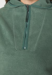Pullover verde in pile con mezza zip, dotato di cappuccio e texture morbida. Dettagli delle cuciture notevoli e colore della cerniera in contrasto.