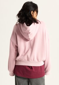 Pink hooded sweatshirt with a relaxed fit and ribbed cuffs, worn over a maroon layered top. Soft texture with a smooth finish.