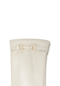 Crème leren laars met een gladde textuur, voorzien van een gouden hardwareaccent en een decoratieve riemdetail bovenaan.