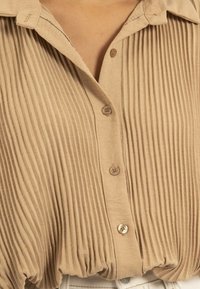 Blouse beige plissée avec un col, dotée d'un devant à boutons et de coutures visibles. Texture douce, ourlet resserré et détails de boutons subtils.