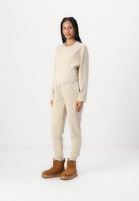 Pull en tricot beige avec taille élastique, associé à un pantalon ajusté assorti. Le mannequin porte des bottines en daim marron avec des semelles robustes.