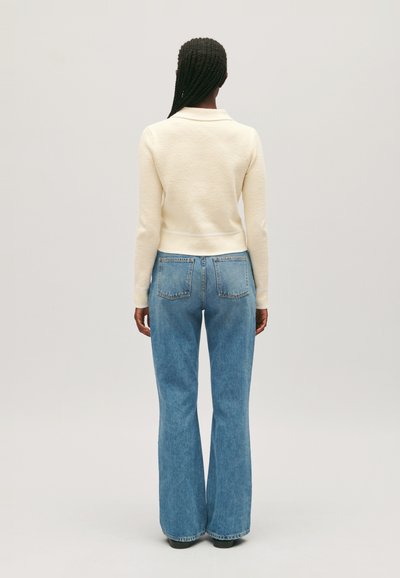 Suéter de punto crema con cintura ajustada, combinado con jeans de denim de tiro alto y corte acampanado en color azul claro, que cuentan con bolsillos traseros. Fondo neutro.