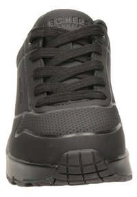 Zapatillas deportivas negras con una parte superior de sintético liso, perforaciones para transpirabilidad, cordones planos y una suela de goma texturizada para agarre.