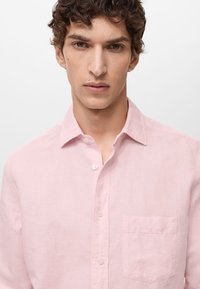 Lichtroze button-up shirt gemaakt van een gestructureerde stof, met een puntige kraag en een borstzak aan de linkerkant.