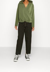 Persona vistiendo una sudadera envolvente verde oliva, pantalones negros con cordones y zapatillas de caña alta en blanco y negro, de pie contra un fondo blanco plano.