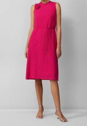 Robe fuchsia sans manches, longueur genou, en tissu texturé, avec une ceinture à la taille, un ornement en forme de rose sur l'épaule, portée avec des sandales à talons argentées.