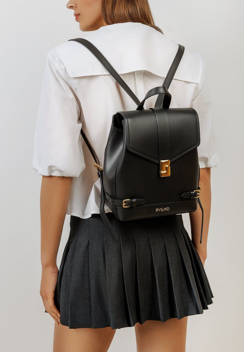 Zaino in pelle nera con un design strutturato, dettagli in hardware dorato e spallacci regolabili. Presenta una chiusura a patta e un dettaglio con logo.