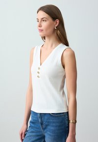 Witte geribbelde mouwloze top met een V-hals, voorzien van drie goudkleurige knopen aan de voorkant. Gecombineerd met lichtblauwe denim jeans.