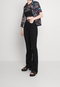 Svarta utsvängda jeans kombinerade med en kort svart topp och en kortärmad, färgglad mönstrad skjorta med neongrafik och vågiga kanter.