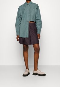 Camicia a righe color teal con design corto, abbinata a una gonna a campana a quadri scuri e scarpe beige chiare con lacci.