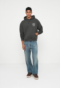 Sötét szürke pamut kapucnis pulóver, kenguruzsebbel és húzózsinórral, részletes grafikával. Világoskék farmernadrággal és barna cipővel párosítva.