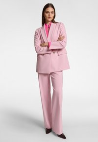 Completo sartorial rosa chiaro con giacca doppiopetto e pantaloni a gamba larga. La giacca ha bottoni dorati; sotto indossa una blusa fucsia.