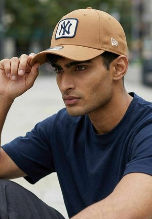 Junger Mann trägt eine hellbraune NY-Baseballkappe und ein dunkelblaues Hemd, richtet die Kappe gerade und schaut leicht seitlich im Freien.