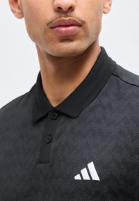 Κοντινή λήψη ενός ατόμου που φοράει μαύρο πόλο με μοτίβο, μαύρα κουμπιά και λευκό λογότυπο Adidas στο στήθος.