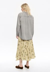 Grijze denim shirt met een klassieke kraag, lange mouwen, achterplooien, gecombineerd met een gele bloemenmaxi-jurk en bruine instapklompen.