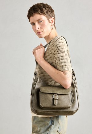 Borsa a tracolla in pelle verde oliva con pattina frontale, tracolla regolabile e dettagli in metallo argentato, mostrata su un modello che indossa un maglione lavorato a maglia.