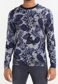 Mann in einem grauen Langarmpullover mit dunkelblauen tropischen Blatt- und Ananasmustern, kombiniert mit dunklen Hosen.