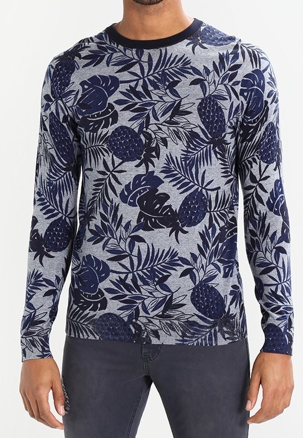 Mann in einem grauen Langarmpullover mit dunkelblauen tropischen Blatt- und Ananasmustern, kombiniert mit dunklen Hosen.