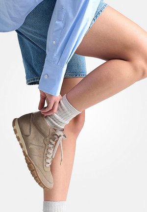 Sneaker alta beige in suede con suola, lacci e logo marroni chiari. Indossato con calze bianche a coste e pantaloni shorts di denim, su uno sfondo chiaro.