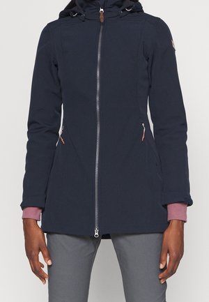 Femme portant une veste à capuche bleu marine zippée avec des poches latérales zippées, superposée sur un haut mauve à manches longues et un pantalon gris.