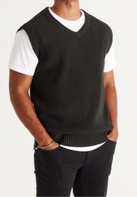 Gilet lavorato a maglia verde scuro con scollatura a V e texture a coste, indossato sopra una maglietta bianca, abbinato a pantaloni neri. Design senza maniche.