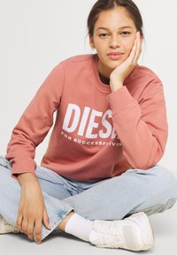 Rosa sweatshirt mit weißem Druck "DIESEL FOR SUCCESS LIVING", kombiniert mit hellblauen Jeans und weißen Sneakers, im Schneidersitz sitzend.