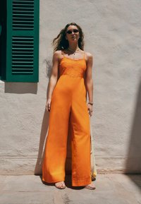 Combinaison sans manches orange avec un corsage brodé de fleurs et un design à jambes larges ; le mannequin porte de petites lunettes de soleil et se tient devant un mur texturé.