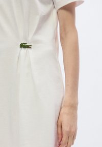 Λευκό βαμβακερό φόρεμα με σούρες στο πλάι και πράσινη κεντημένη λεπτομέρεια κροκόδειλου. Κοντά μανίκια και άνετη εφαρμογή.