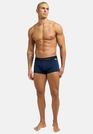 Spierkrachtige man die blootsvoets staat, gekleed in marineblauwe boxershorts met zwarte tailleband, tattoo op de linkervoorarm, kijkt naar links tegen een witte achtergrond.