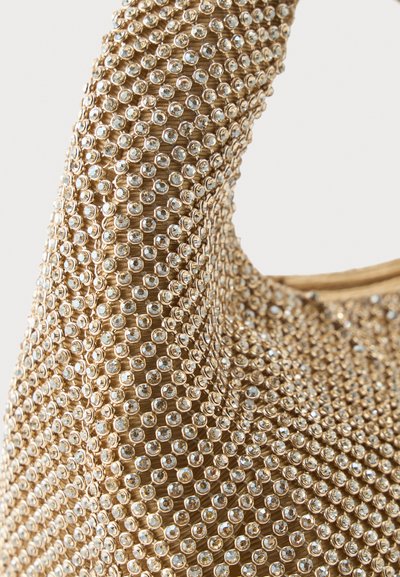 Približan posnetek traku za torbico iz bež tkanine, goste prekrite z majhnimi prozornimi kristali, razporejenimi v enakomeren vzorec.