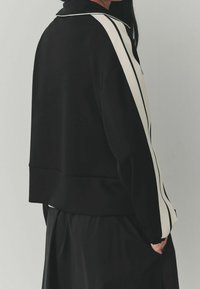 Chaqueta deportiva negra con rayas blancas en las mangas, con cuello y un ajuste holgado. Textura suave, con un diseño ligeramente recortado.