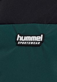 Tejido acolchado verde y negro con un parche de goma en blanco y negro que dice "hummel SPORTSWEAR". Textura suave, diseño deportivo.