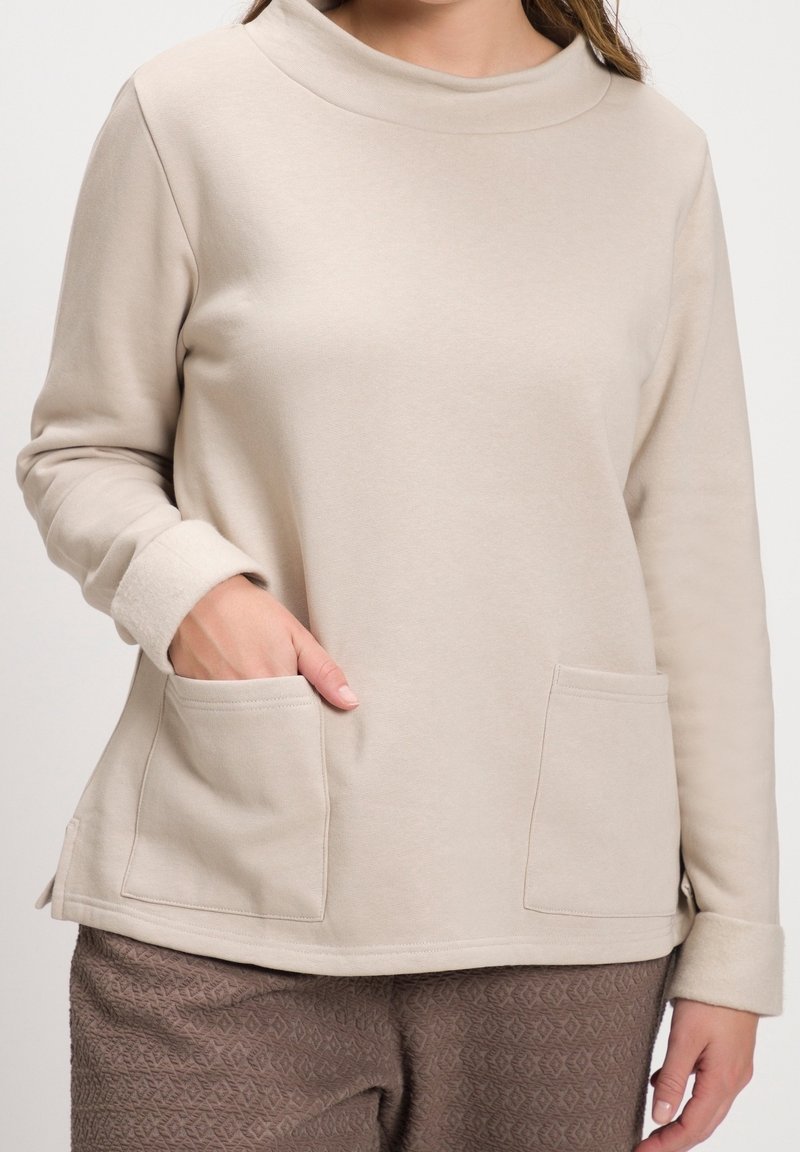 Femme portant un haut beige à manches longues avec deux poches devant et un pantalon marron texturé, main placée dans la poche gauche.