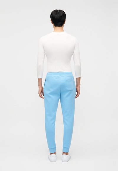 Άνδρας που φοράει εφαρμοστό άσπρο πουκάμισο με μακριά μανίκια, ανοιχτό μπλε παντελόνι τύπου jogger με πίσω τσέπη και άσπρα αθλητικά παπούτσια, στέκεται πλάτη σε απλό φόντο.