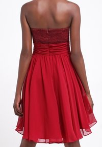 Robe bustier en ligne A de couleur rouge profond. Corsage en dentelle froncé, jupe fluide en chiffon, ourlet à hauteur du genou. Fermeture éclair au dos visible.